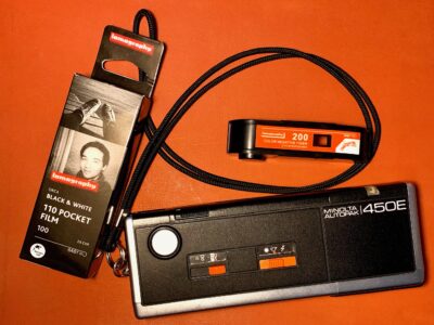 A Minolta 110 film camera with a color film cassette and a box of lomography Black and white film next to it.