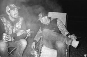 Two men smoking cigars by a smokey fire in black and white.