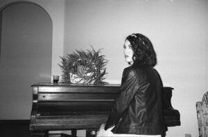 Black and white woman in a dress sitting by a piano looking over her shoulder.