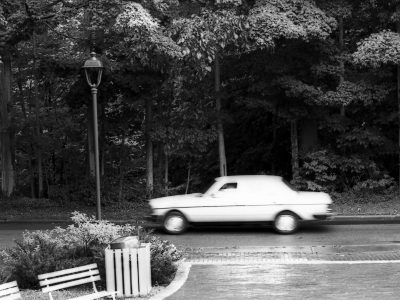 A Black and White film photo of a car driving on a road in the rain. The car is moving quickly and appears blurry.