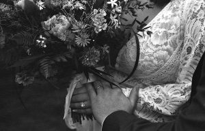 Black and white film photo close up of wedding flowers and a wedding dress and a couple holding hands with their rings visible taken on a Canon AE-1.
