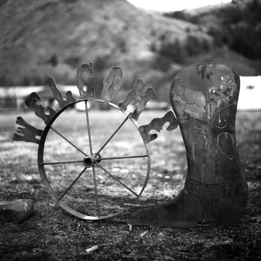 A BW photo of a metal welcome sign on a wheel with a metal cowboy boot shape on the right side.